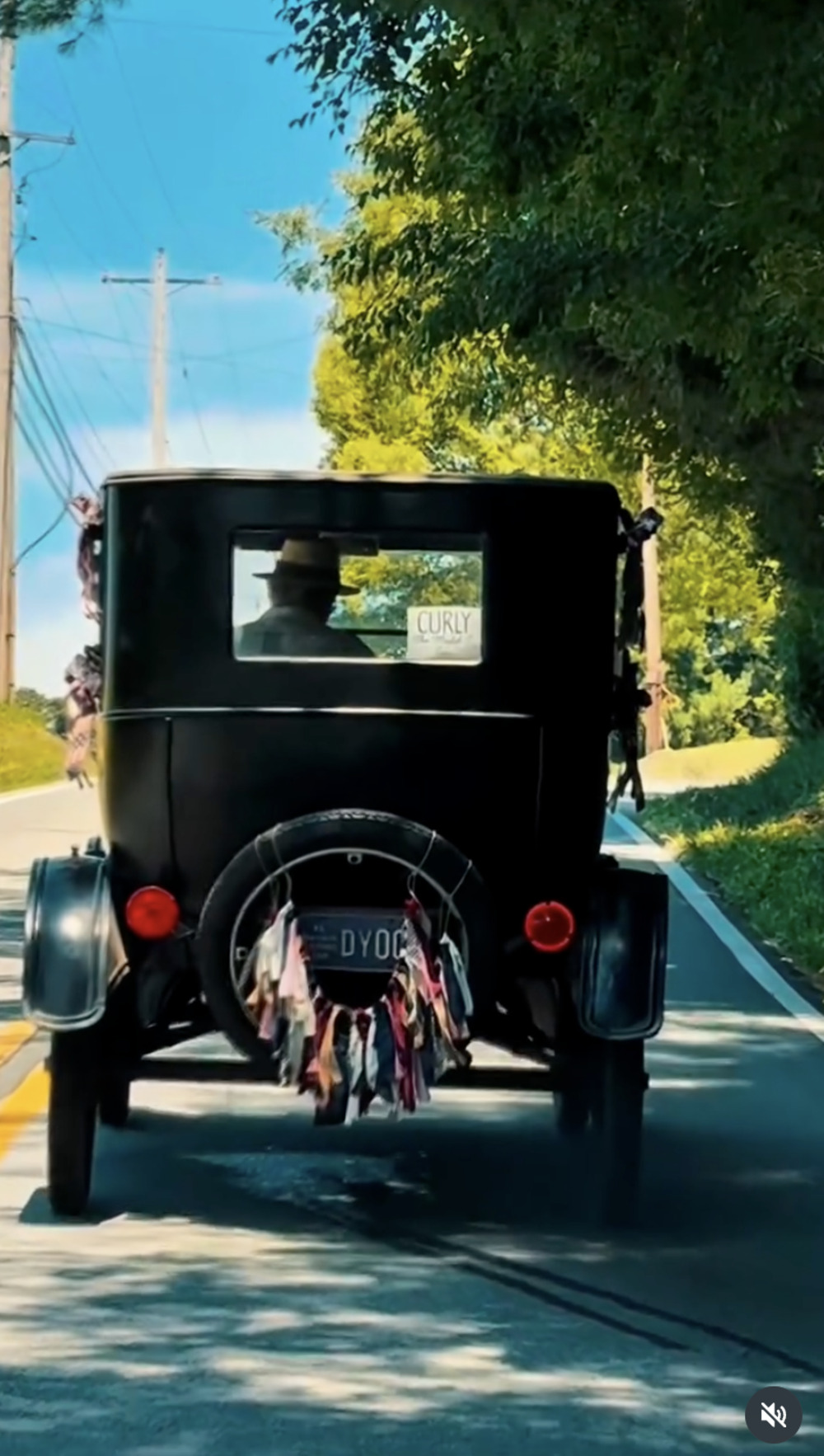 Curly, a black 1923 Model T sedan, driving away on a sunny two-lane road with ribbons on the spare tire and a Curly sign in the rear window